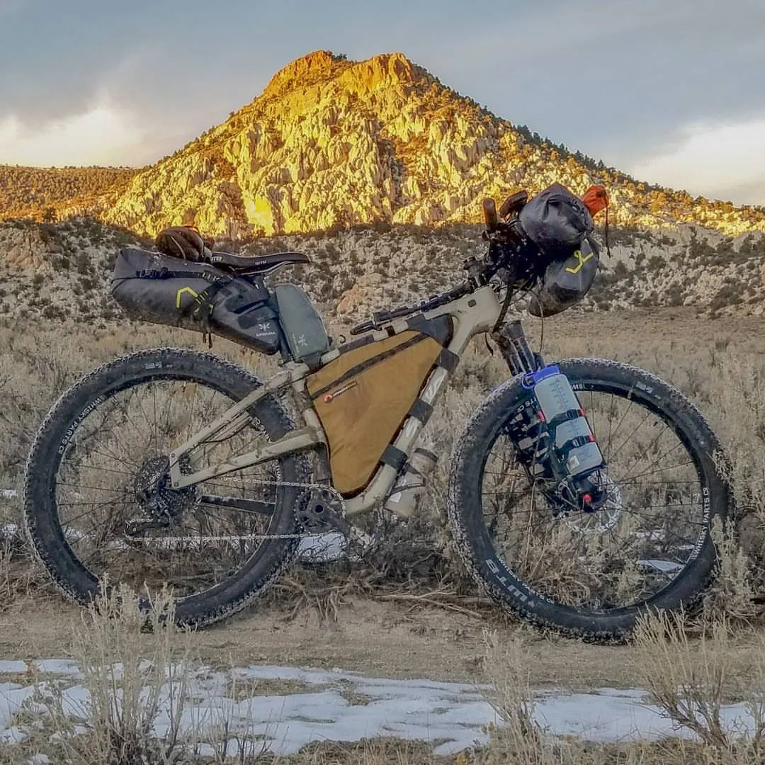 A loaded bicycle stands in pale grass beneath a sunlit rocky hillside.