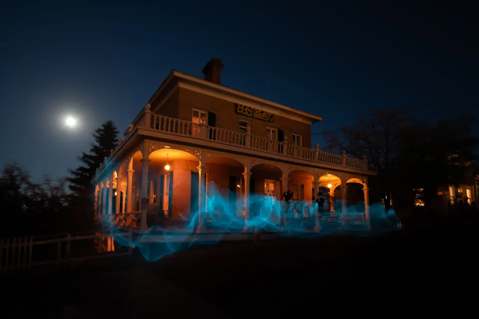 Blue light trails gather around a warmly lit timber house beneath the moon.