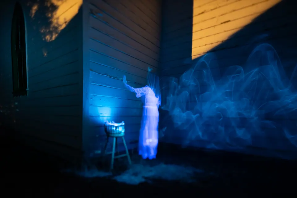 A blue light-painted figure and luminous wisps hover beside the wall of a dark wooden building.