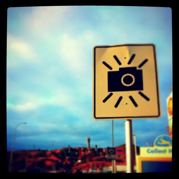 A black camera symbol appears on a yellow roadside sign beneath a blue sky.