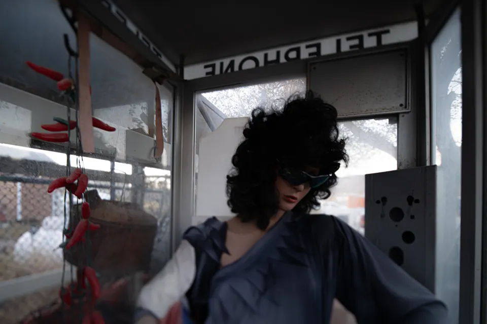 A mannequin in dark glasses occupies a weathered telephone booth.