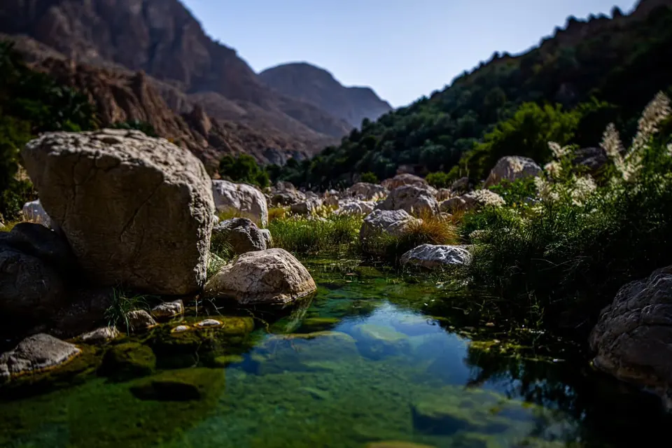 A clear stream winds between boulders and green vegetation below mountain walls.