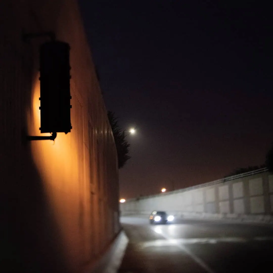 A wall lamp and distant headlights illuminate a narrow passage at night.