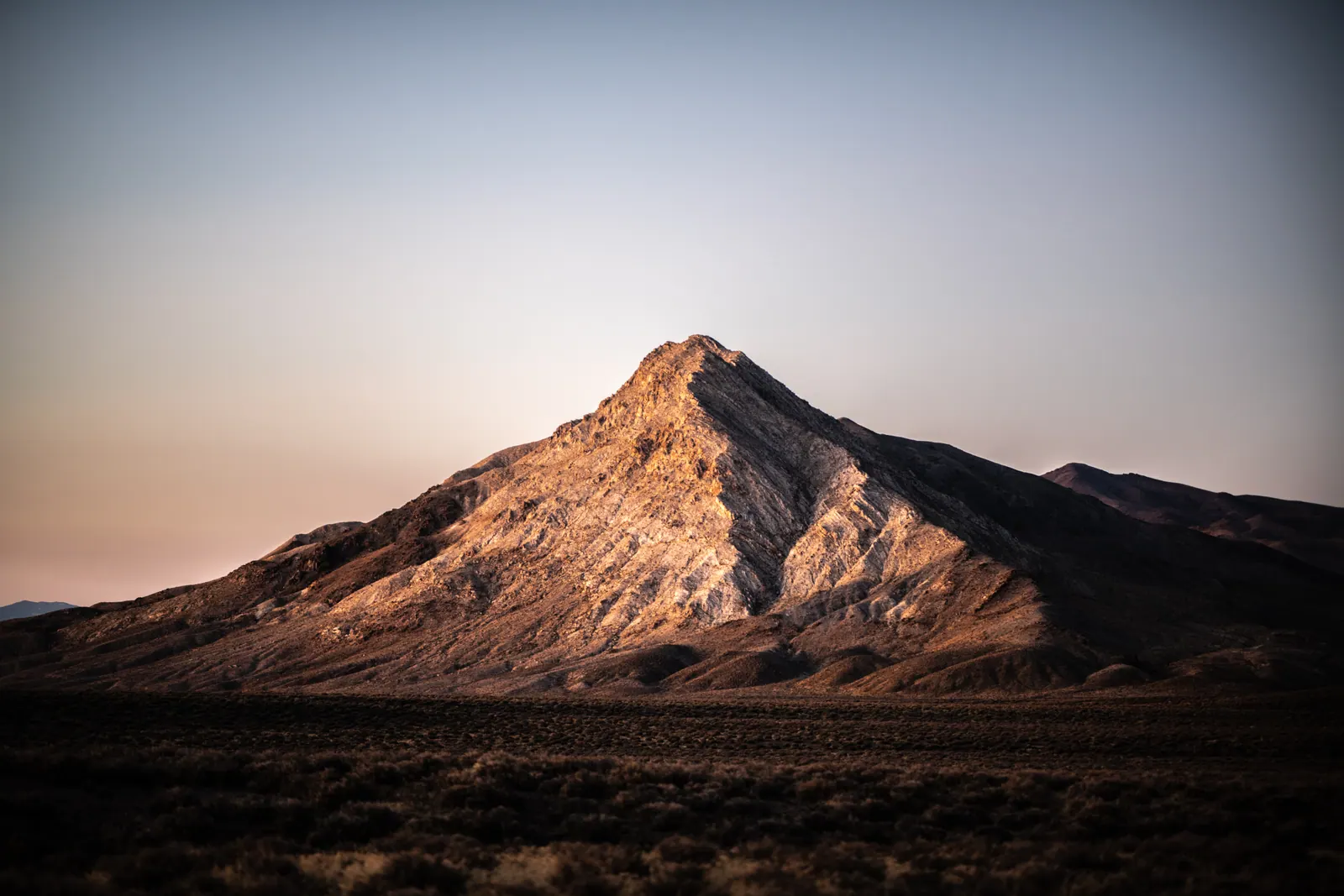 The face of a solitary mountain glows above a dark desert plain.