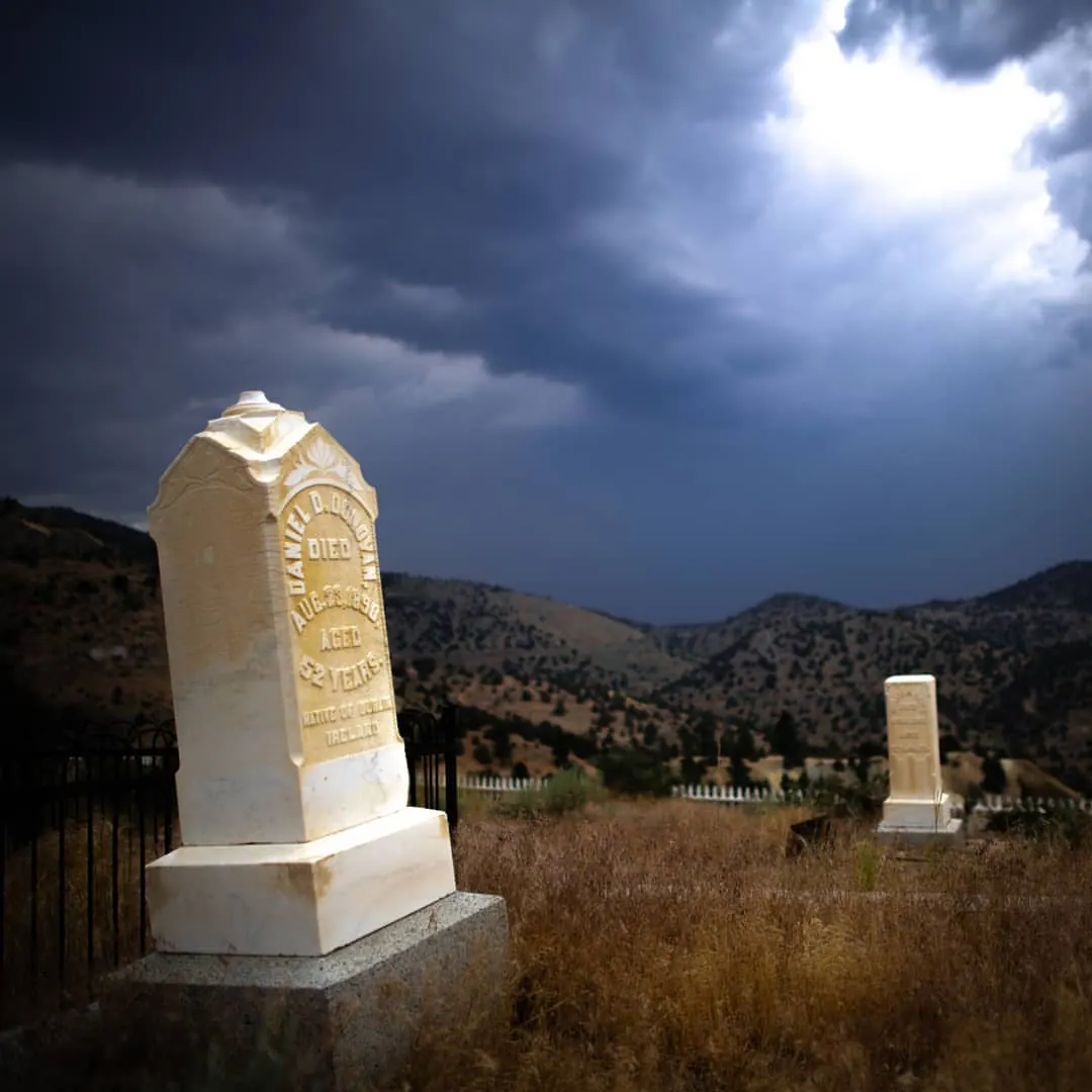 A pale grave marker stands on a hillside beneath a dramatic dark sky.