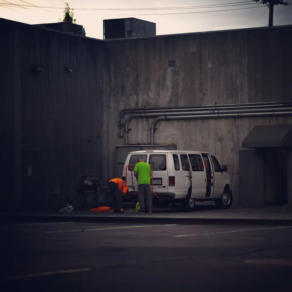 A person stands beside a white van near a dark industrial wall.