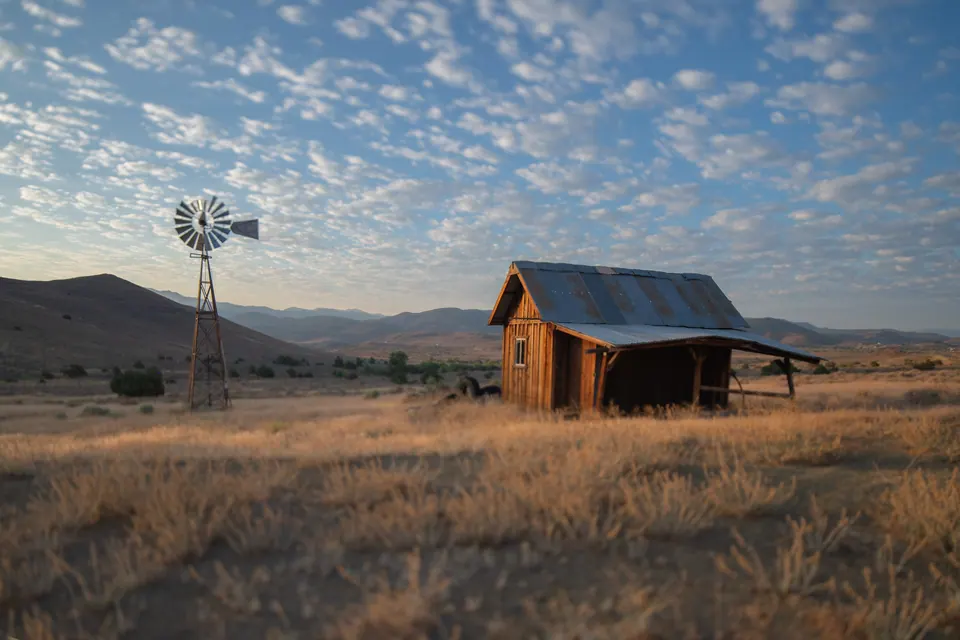 A small timber cabin and a windmill stand in golden grass below mountains and a patterned sky.