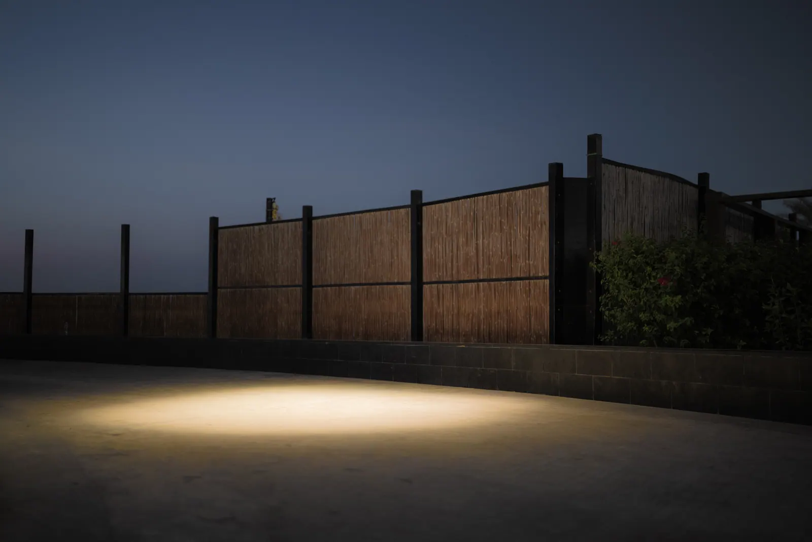 A single pool of warm light illuminates an empty paved space beside a tall fence.