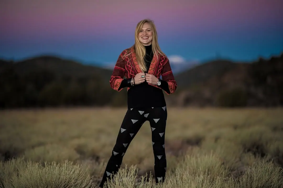 A person in a red jacket stands in an open field beneath a pink and blue evening sky.