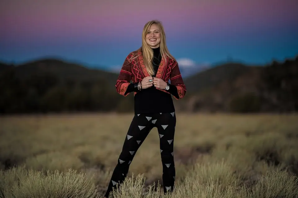A person in a red jacket stands in an open field beneath a pink and blue evening sky.
