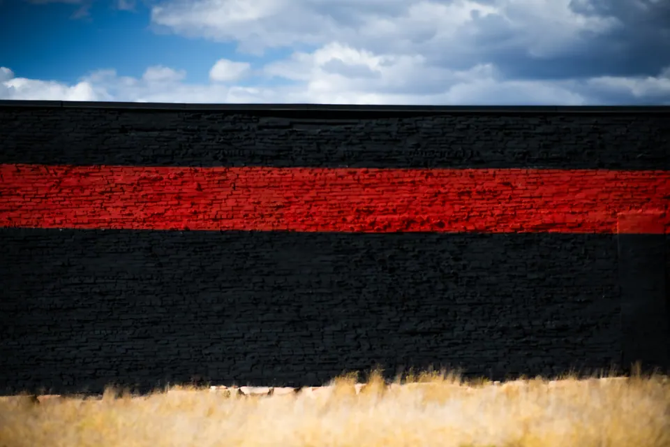 A single red stripe cuts across a dark wall beneath a blue, clouded sky.