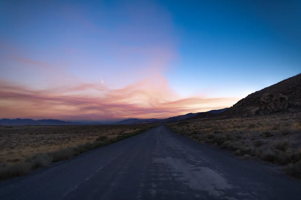 A narrow empty road runs toward distant mountains beneath a pink and blue sky.