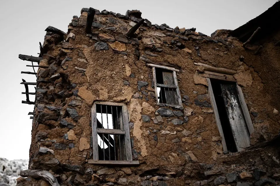 A broken window and exposed beams remain in a crumbling stone house.