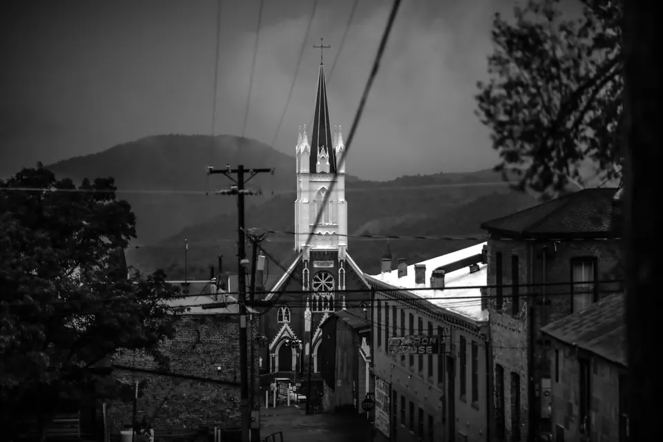 A church steeple rises between buildings and overhead wires on a hillside street.