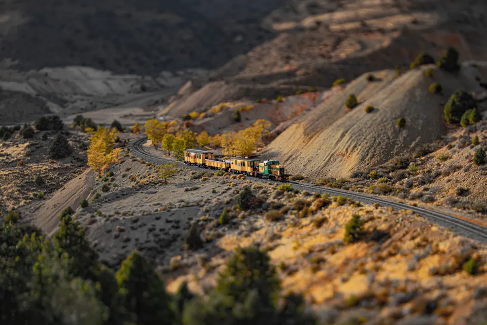 A small train follows a track cut into a dry, sunlit hillside.