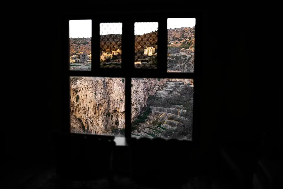 A dark window frame divides a sunlit view of a rocky village.