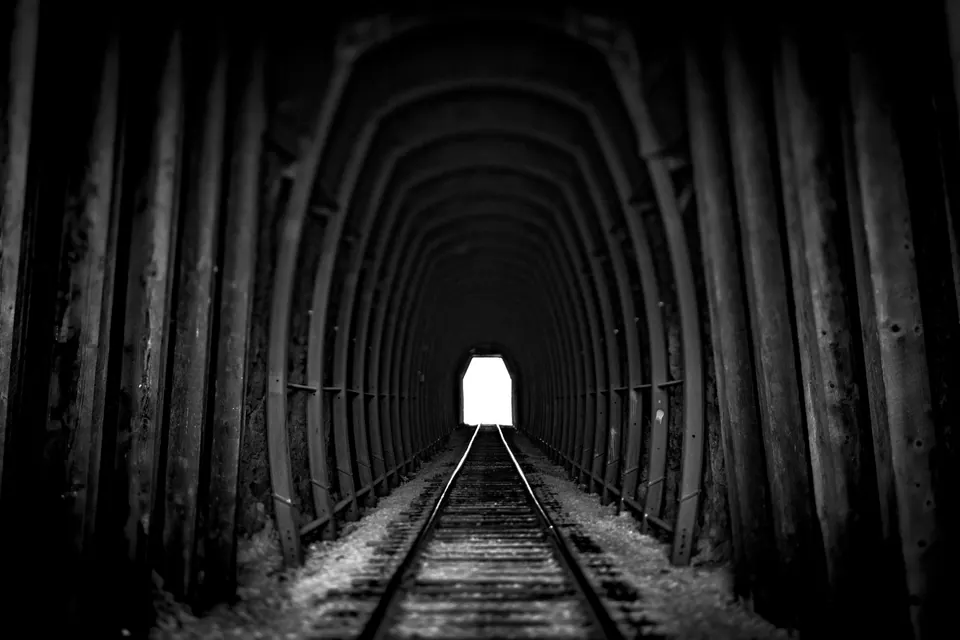 Repeating timber arches and railway tracks lead toward a white opening at the end of a dark tunnel.