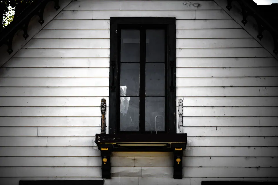 A single dark window sits in a white clapboard wall below a pitched roof.