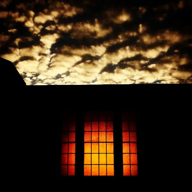 An orange-lit window appears beneath a dark sky filled with textured clouds.