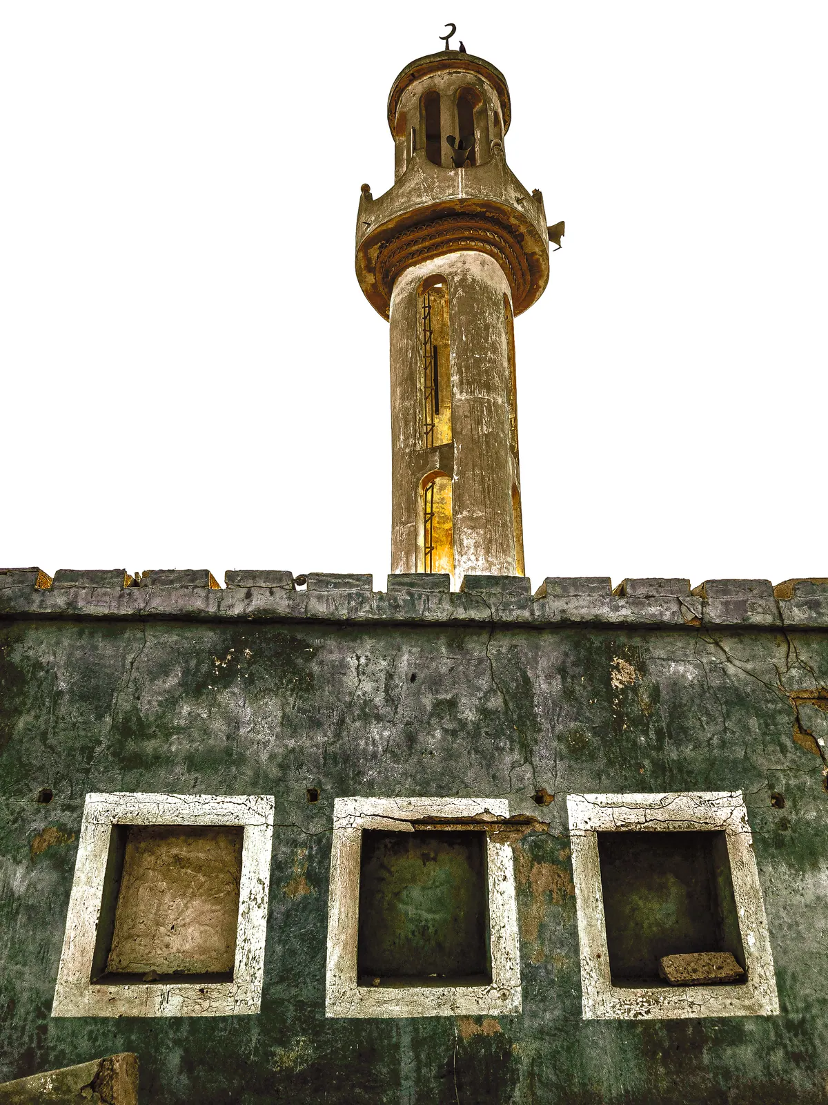 A slender minaret rises above a weathered green wall punctuated by pale window frames.