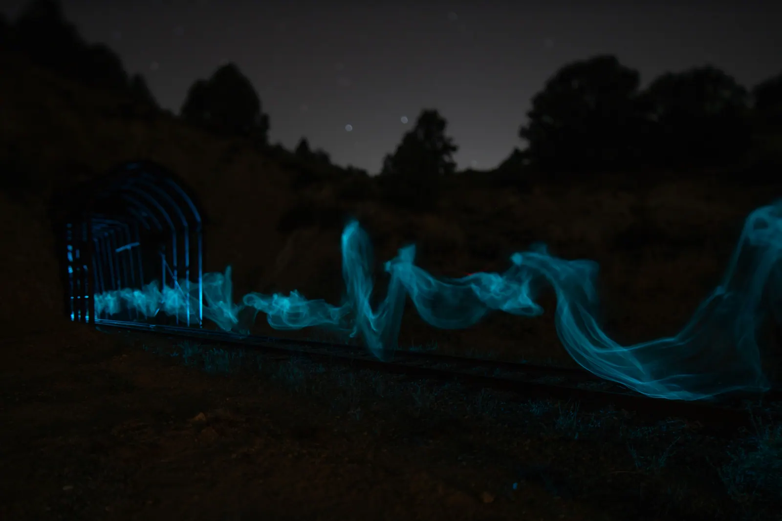 Pale blue light curls across dark ground beneath a starry sky and silhouetted trees.