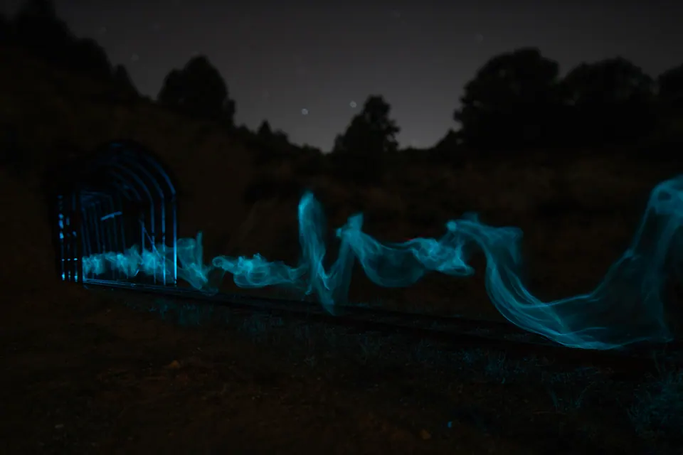 Pale blue light curls across dark ground beneath a starry sky and silhouetted trees.