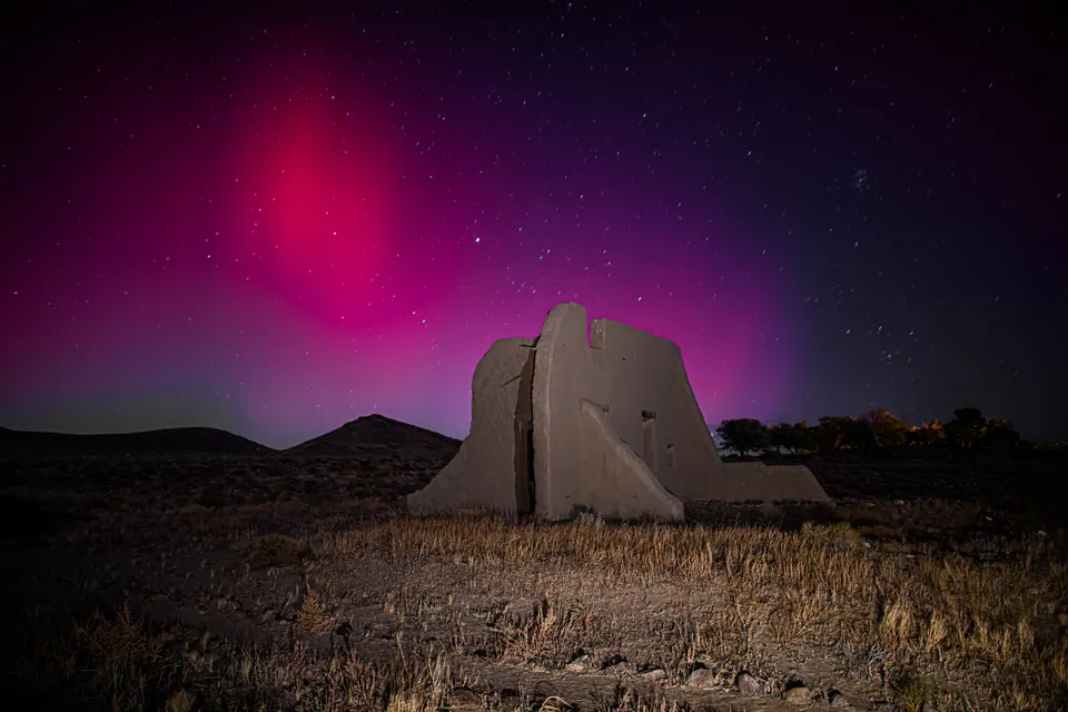Violet and pink light fills the night sky above the remains of an earthen building.