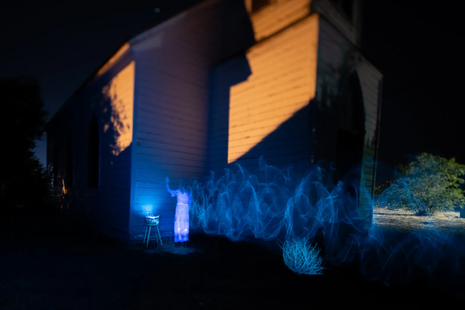 A trail of blue light drifts beside a wooden house lit by warm yellow light.