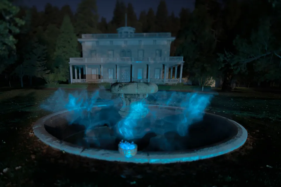 Blue light drifts above a circular fountain in front of a dark mansion.