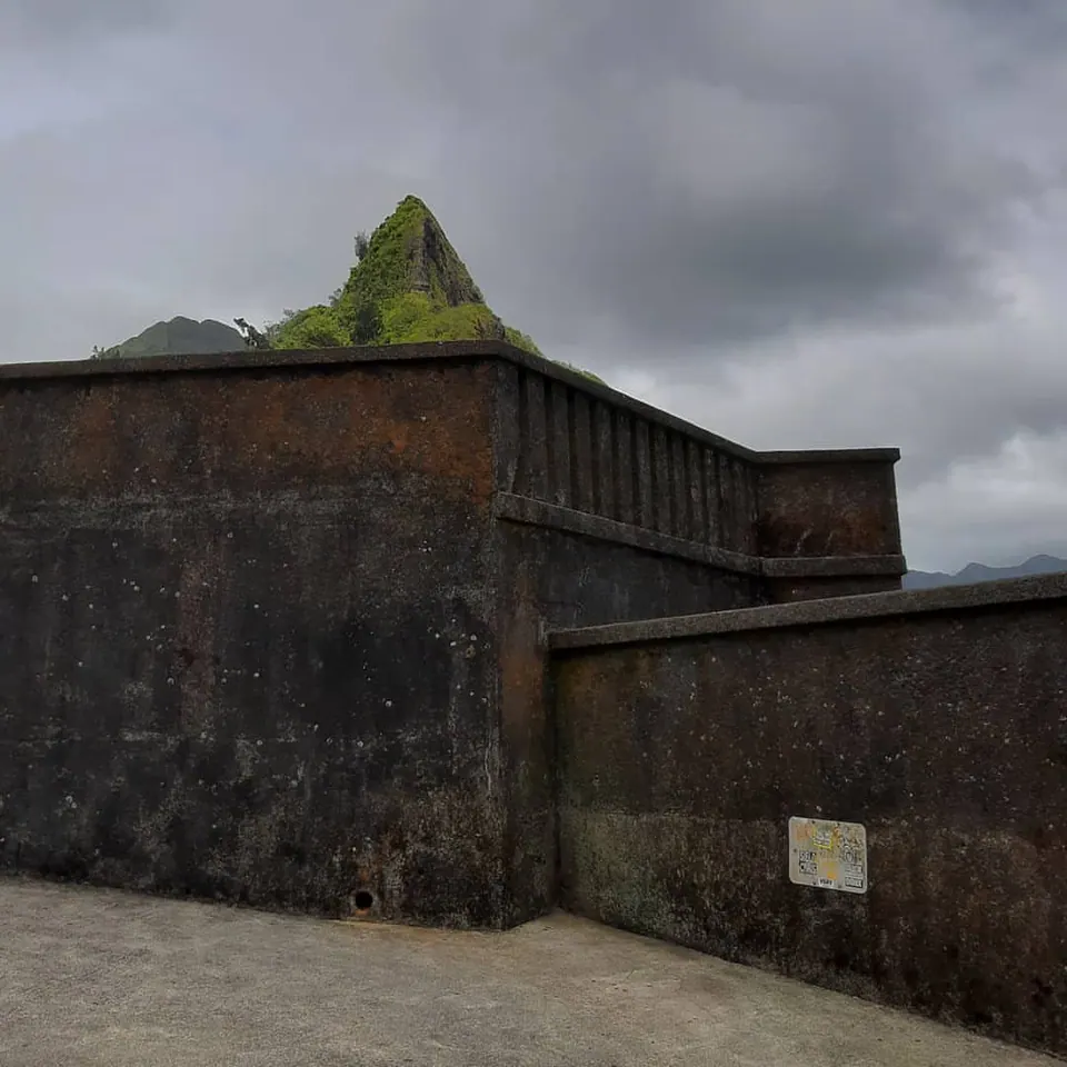 Angular concrete walls stand beneath a pointed green mountain and grey clouds.