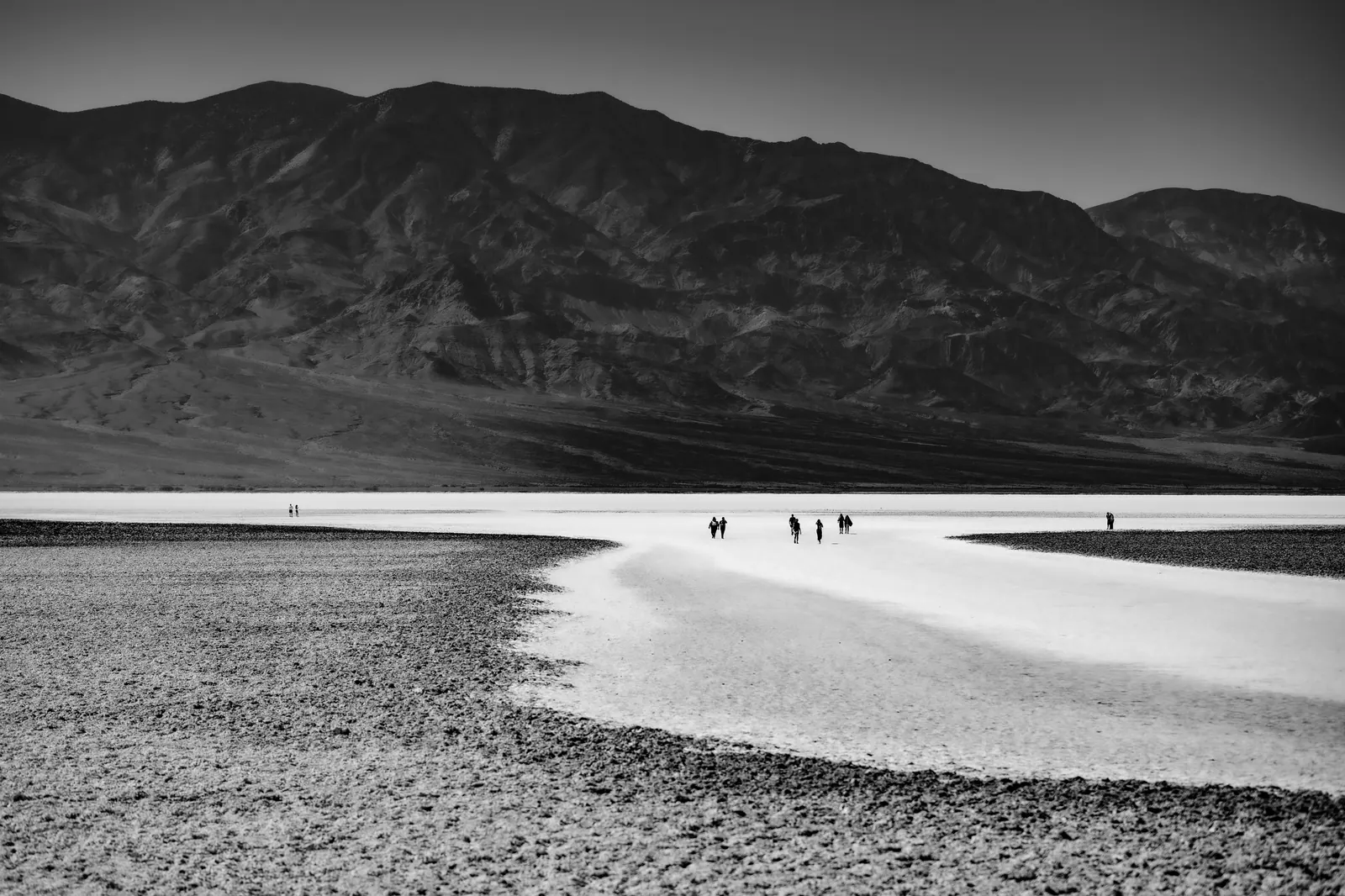A few distant people cross a pale salt flat beneath dark mountains.
