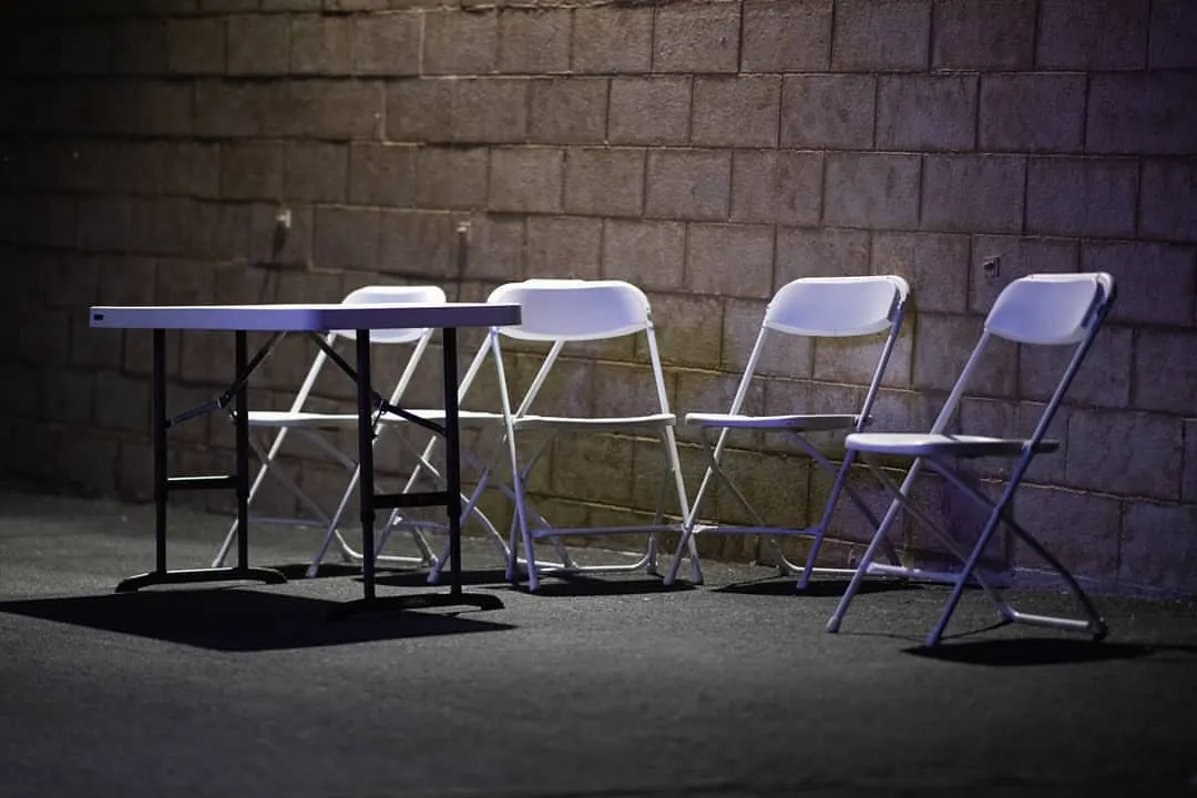Pale folding chairs and a small table sit against a dark brick wall.