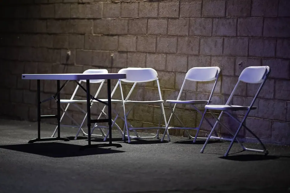 Pale folding chairs and a small table sit against a dark brick wall.