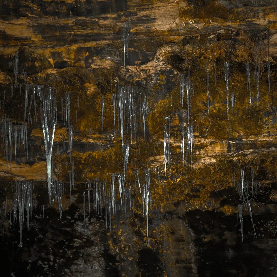 Long icicles hang beneath a layered rock face reflected in dark water.