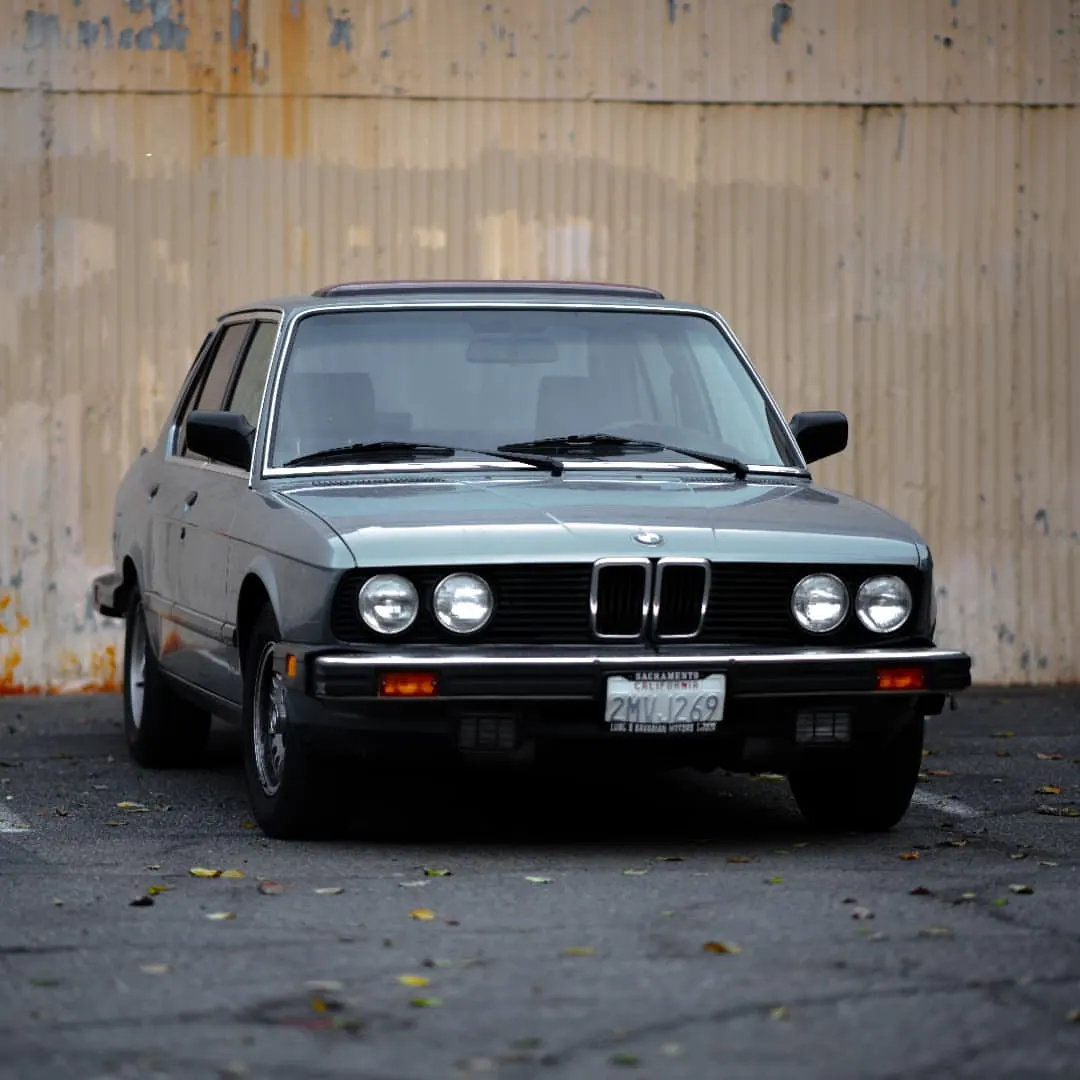 A small grey car stands against the weathered wall of an industrial building.