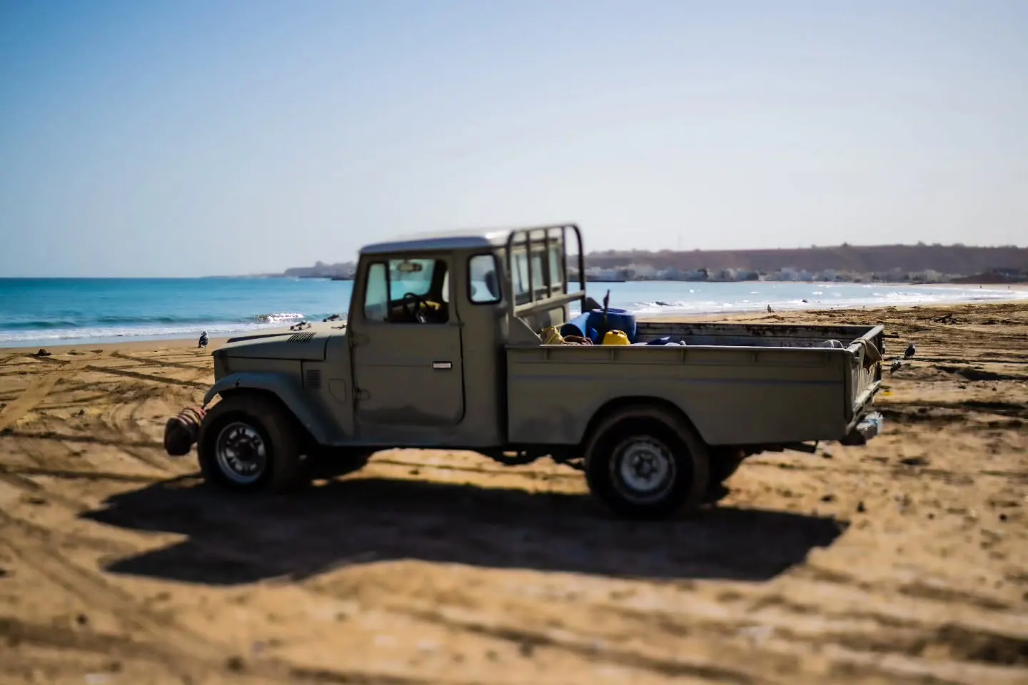 A small pickup truck stands on sandy ground beside a turquoise coast.