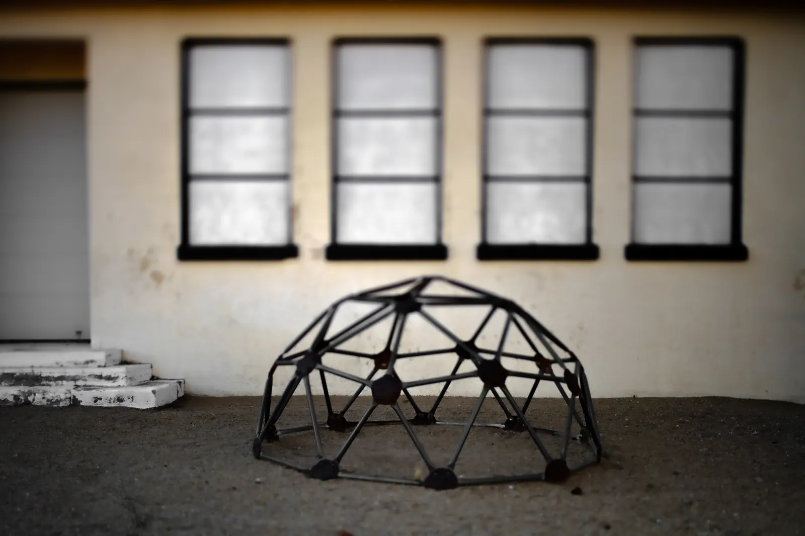 A geometric climbing frame sits in front of a weathered building with a row of dark windows.