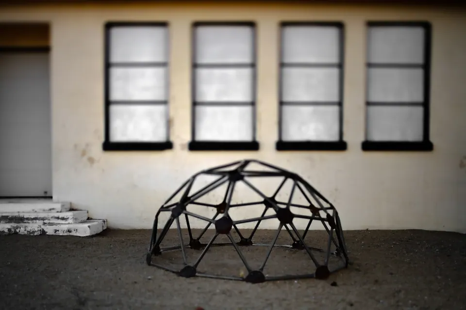 A geometric climbing frame sits in front of a weathered building with a row of dark windows.