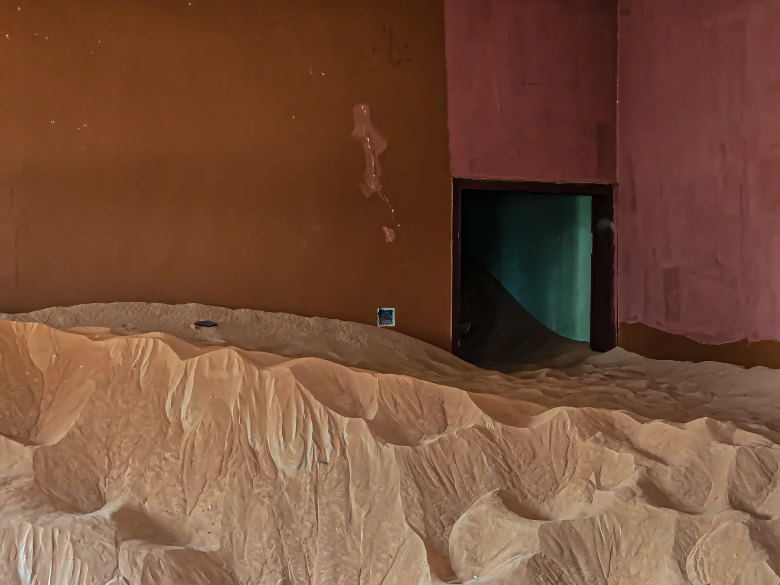 Wind-shaped sand gathers against a red and ochre wall with a small green doorway.