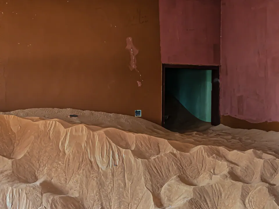 Wind-shaped sand gathers against a red and ochre wall with a small green doorway.