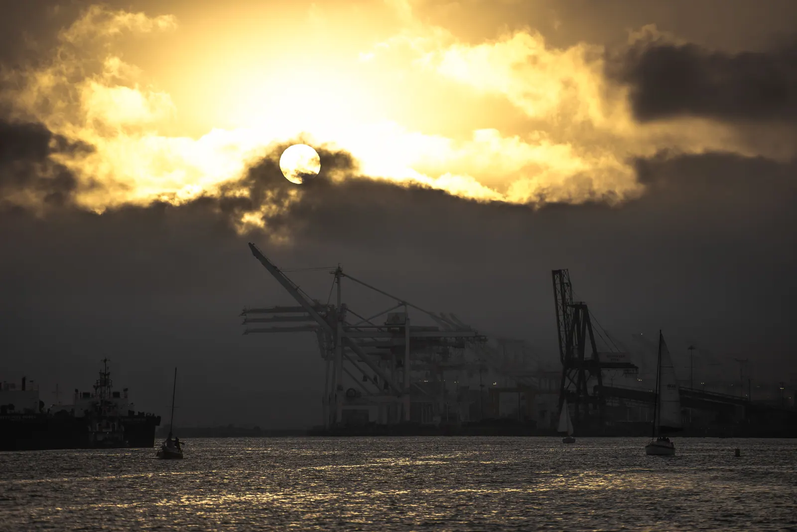 Bright sunlight breaks through heavy clouds above water and port cranes.