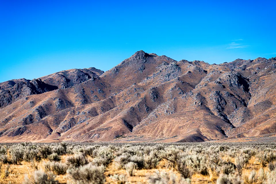 Sharp mountain ridges rise above pale desert shrubs beneath a vivid blue sky.