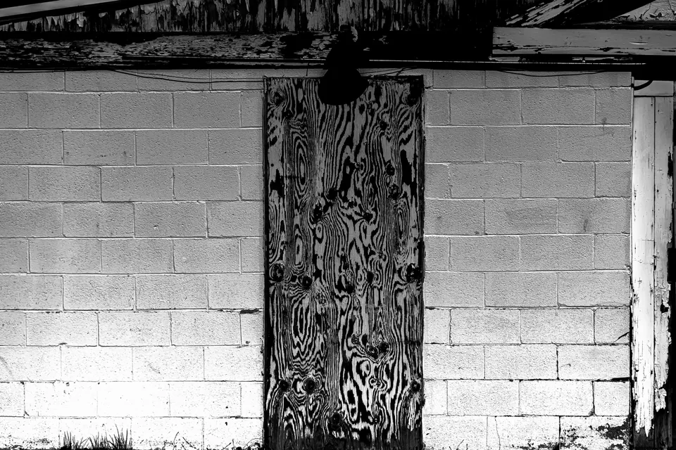 A boarded door interrupts a pale brick wall in a stark monochrome scene.