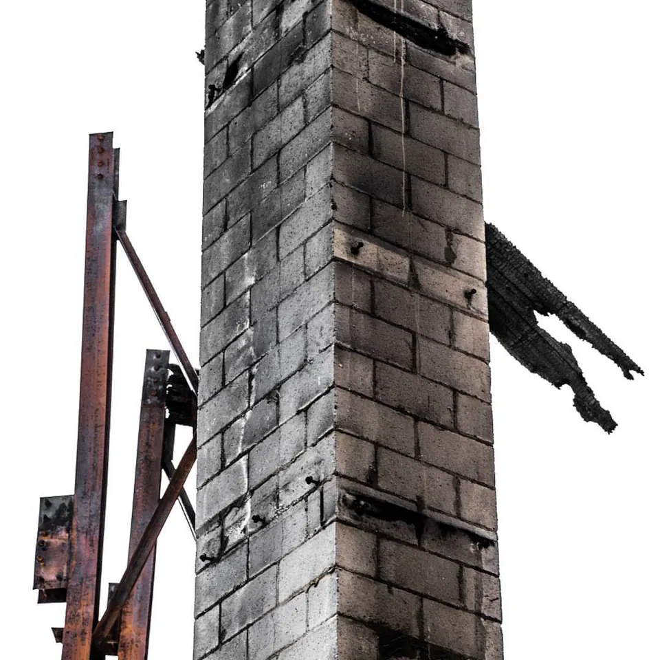 A tall brick chimney and a dark beam remain against a pale sky.