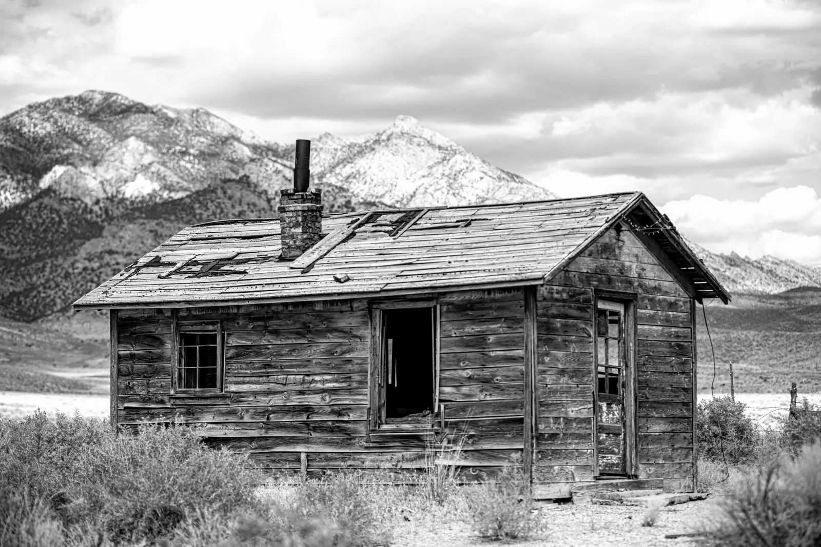 An abandoned wooden cabin faces snow-dusted mountains in black and white.