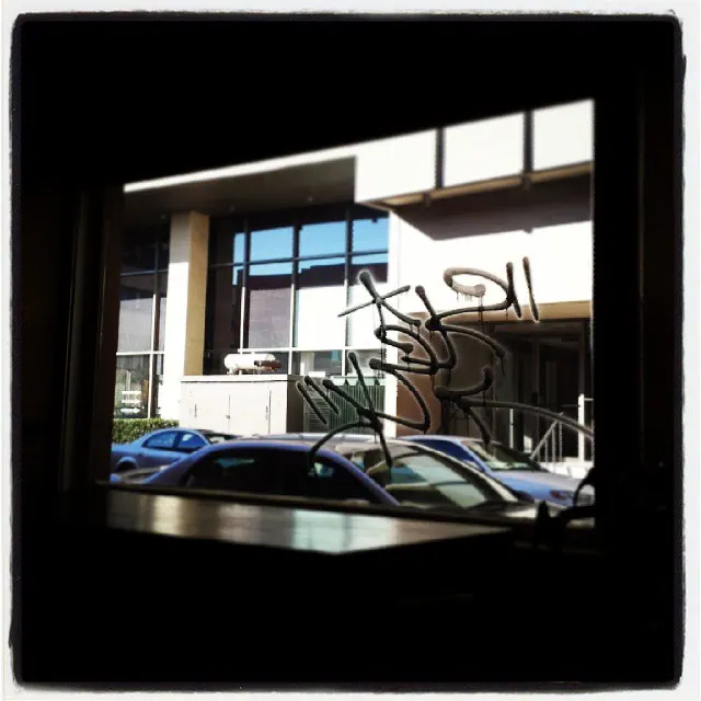 A parked car appears through a dark window bordered by signs and reflections.
