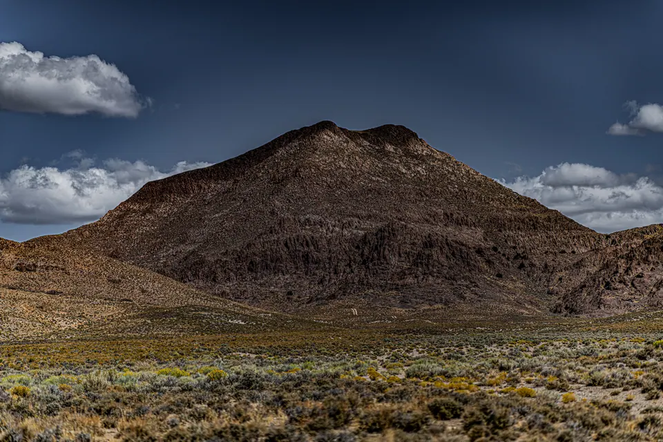 A solitary dark hill rises above pale desert plants under scattered clouds.