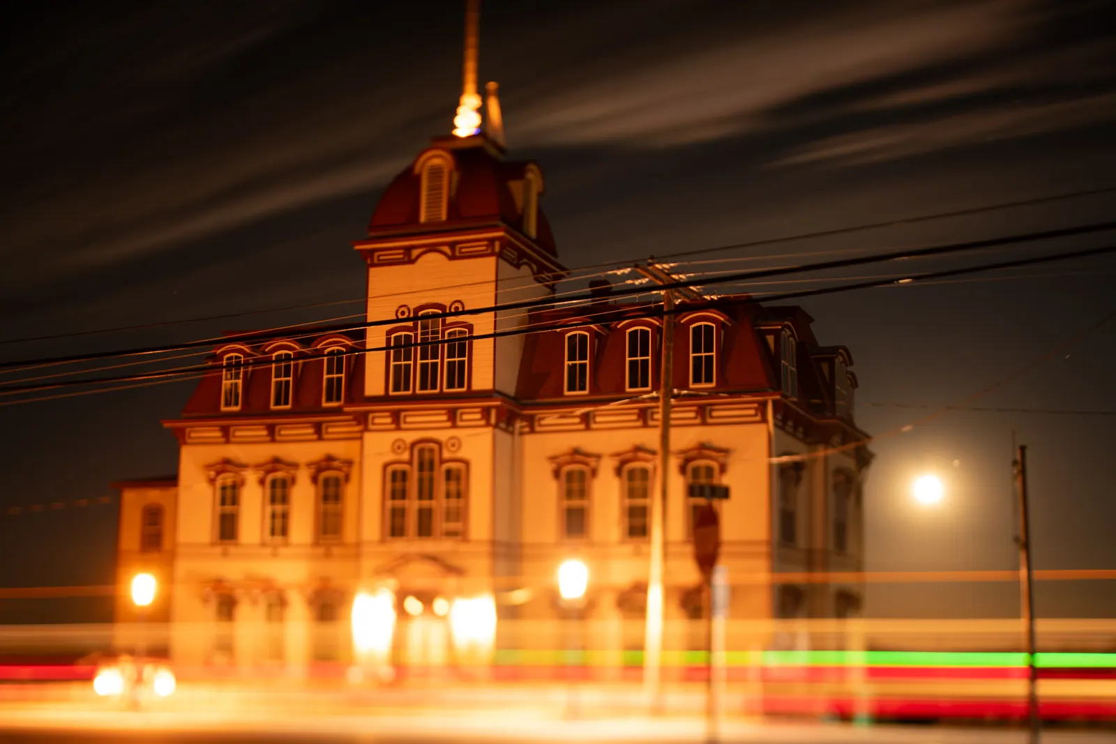 An illuminated historic schoolhouse stands against the night as coloured traffic trails pass.