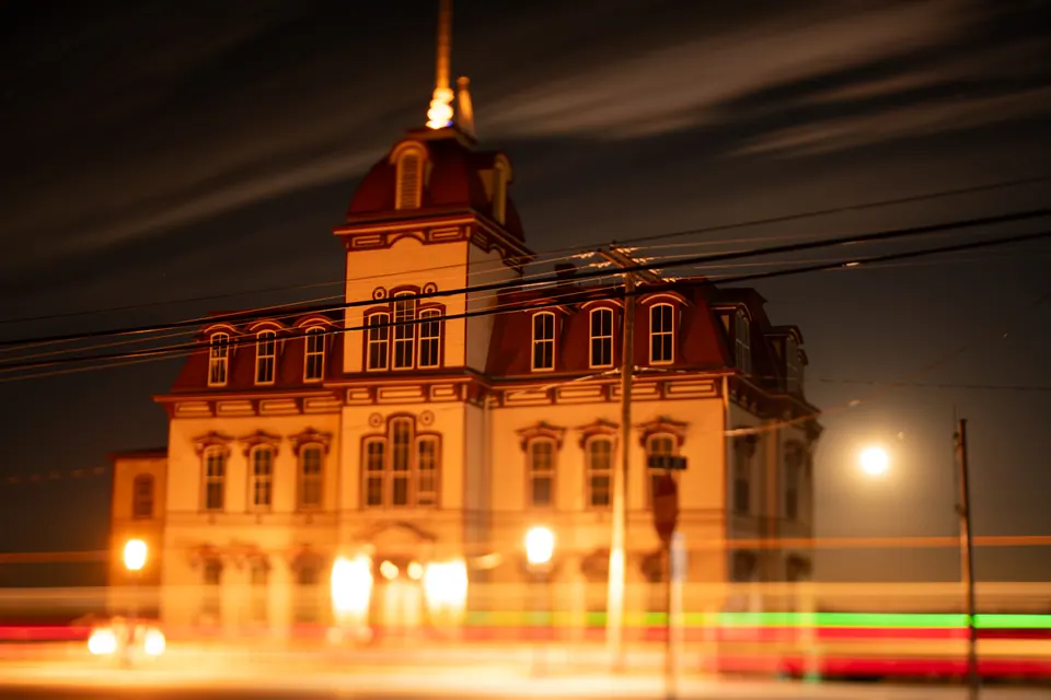 An illuminated historic schoolhouse stands against the night as coloured traffic trails pass.