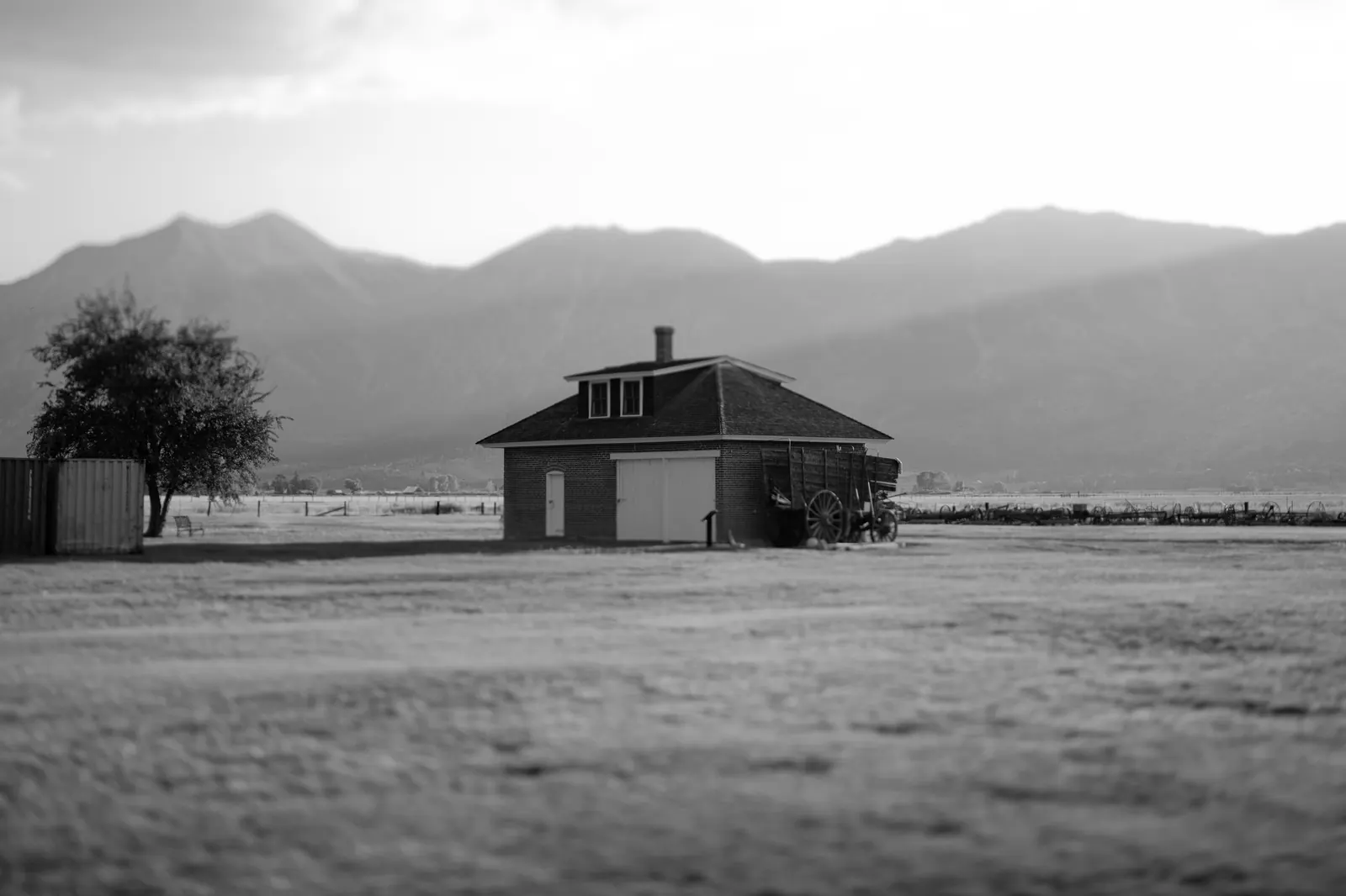 A solitary house sits in an open field below distant mountains in black and white.